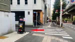 歩道の間の横断歩道　左手にハブの四角い看板、右側は車道　横断歩道上に、赤色で左向きの矢印が表示されている
