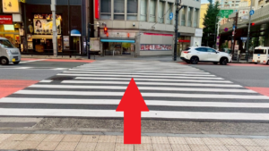 横断歩道　渡った先の正面に交番、左手に水餃子店、右手に三菱ＵＦＪ銀行　赤色で、真っすぐの矢印画像が横断歩道上に表示されている