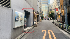 道路　左側のビルの壁に掲示板があり、劇団俳優座の公演ポスターや取材記事などが貼ってある　右手奥に麻布警察署の大きな看板がみえる　ビルの入り口が赤丸で囲まれている　
