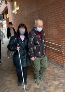 シアタートラムまえで、はくじょうを持った視覚障害の女性を、劇団員のもりはじめが誘導しています