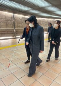 さんげんぢゃや駅で、はくじょうを持った視覚障害の女性を、劇団員のさとうあかりが誘導しています