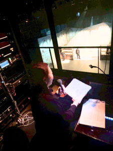 舞台をうえからみおろすことができるオペレーションブースで、はいばらまゆみが、マイクをひだりて、げんこうをみぎてにもち音声ガイドをしています