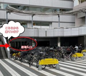 シアターカイのちゅうりんじょうの写真です たくさんの自転車がならべられており、そのおくにげきじょうのいりぐちがみえます ひだりおくにあかいみぎやじるしがかいてあり、こちらからきていますというふきだしがついています