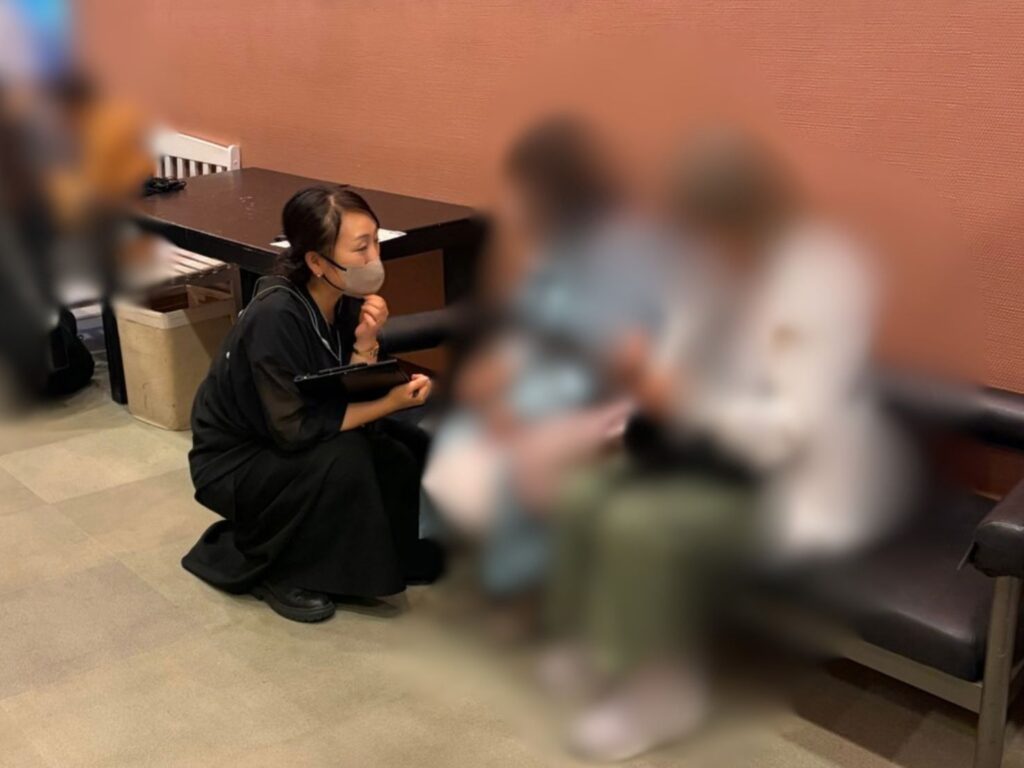 茶色の壁があるロビーで、黒い服を着てマスクを着用したはいばらまゆみがバインダーを持ち、しゃがみ込んでソファに座る2人のりようしゃと向き合っている。はいばらはりようしゃの目線に合わせて姿勢を低くし、耳を傾けながら会話や案内を行っている。