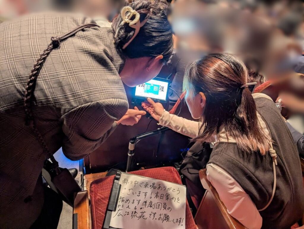 客席内でチェック柄の服を着たひろえるかとこんどうまりあがかがみ込み、文字が表示された小型モニターの画面を一緒に覗き込みながら指で操作している。手前の座席には「雨の中ご来場ありがとうございます!」などの挨拶が手書きされた用紙が置かれている。