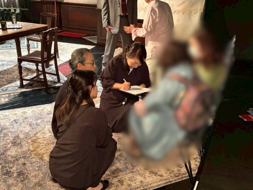 洋間風の舞台セットのうえで、黒い服を着た3人の女性(てまえからますだあかね、あまのまゆみ、はいばらまゆみ)が絨毯の上にしゃがみ込み、はいばらが手元のアンケートにペンで記入を行っている。その傍らにはぼかし処理が施されたりようしゃの姿がある