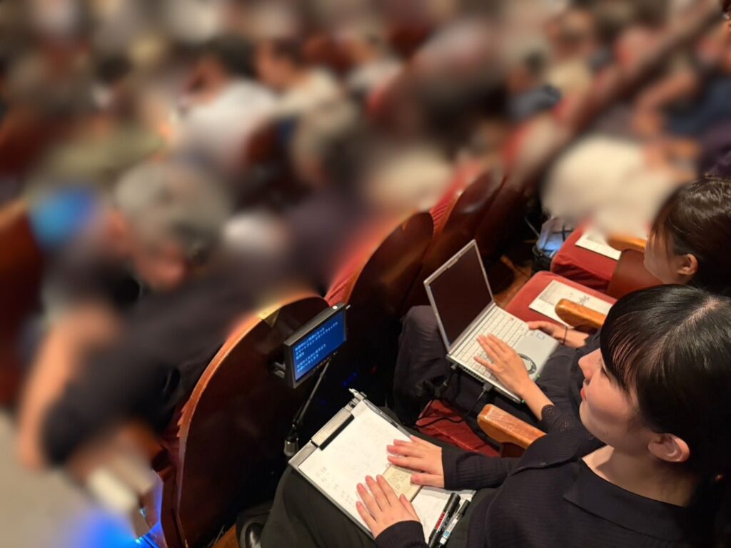 満席の客席で、黒い服を着た2人の女性スタッフが並んで座り操作を行っている。手前の女性(もりなつこ)は膝の上のバインダーに置いた資料に両手を添えており、隣の女性(こんどうまりあ)は膝の上でシルバーのノートパソコンのキーボードを入力している。座席の前方には小さな文字表示用モニターが設置されている。