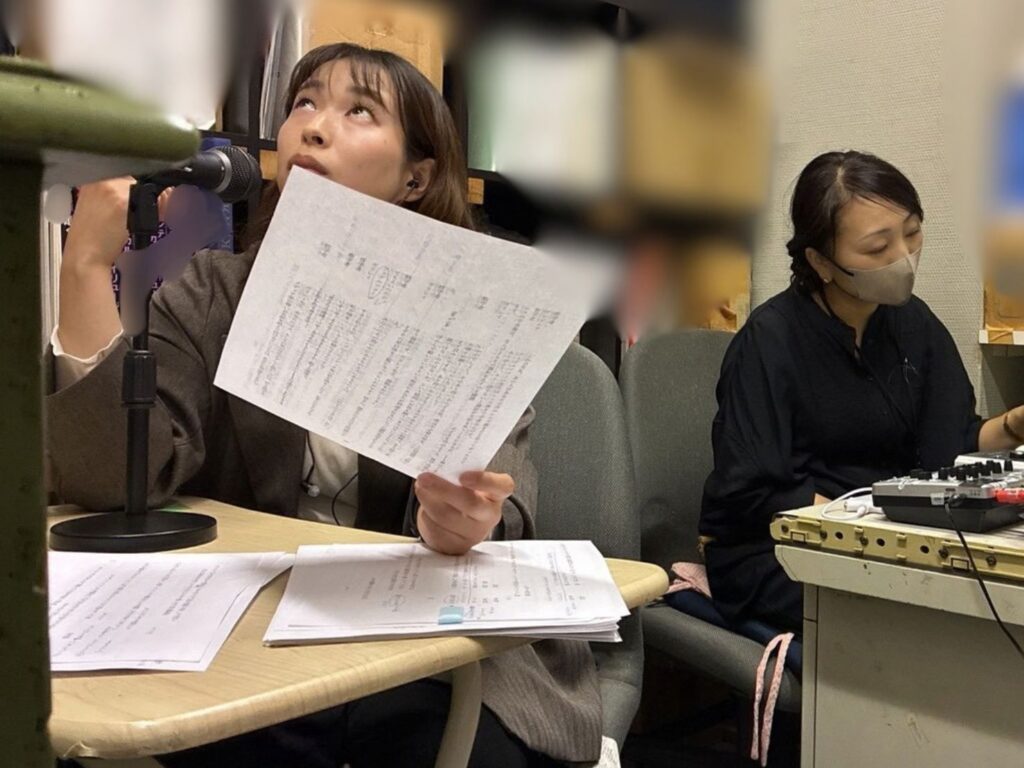 室内で茶色のジャケットを着た、かにさわれいらが手に原稿を持ち、デスクのうえのスタンドマイクに向かって上を見上げながらアナウンスを行っている。その隣のデスクでは、マスクを着用し黒い服を着たはいばらまゆみが機材の前に座り、音響操作を行っている。デスク上には複数の書類が広げられている。