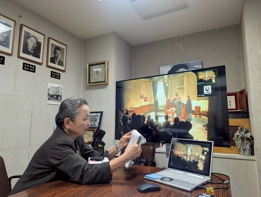 劇団員のあまのまゆみが、マイクに向かい、資料を手にして、演劇が上映されている大きなモニターを見つめている。モニターの画面と、デスク上の開いたノートパソコンの画面には、客席から見た舞台の様子がうつし出されている。