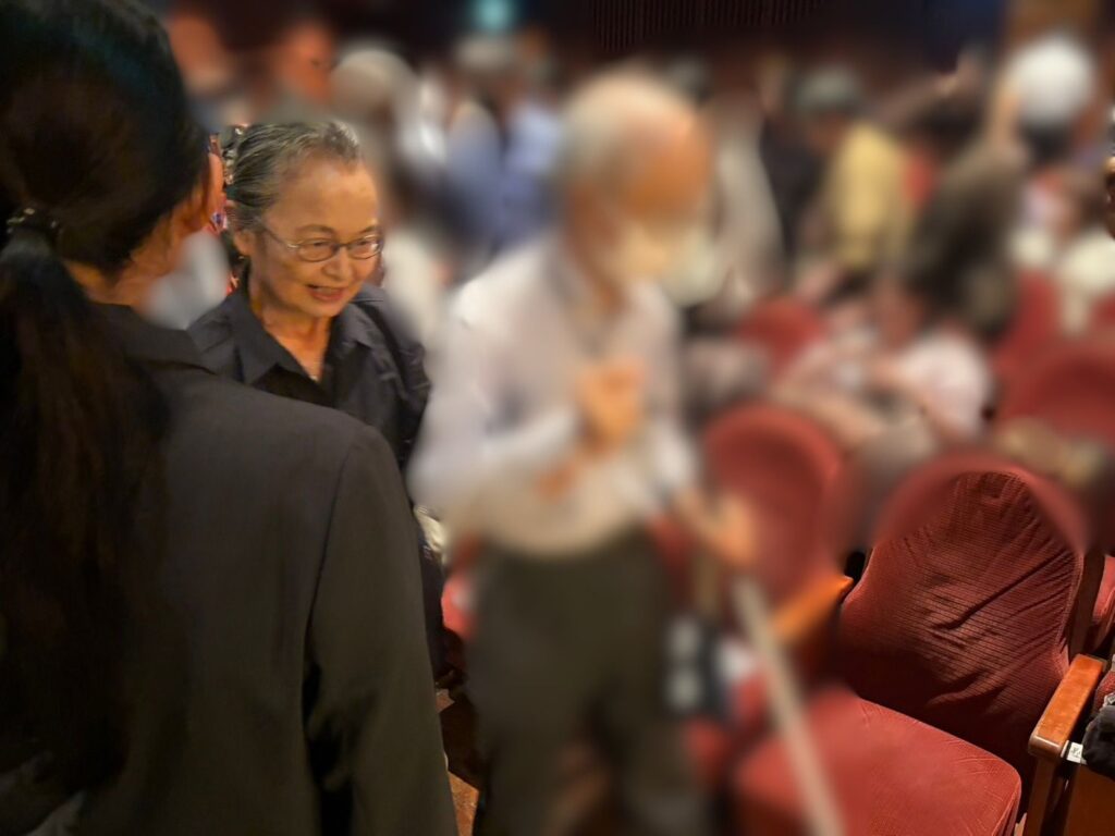多くの観客で賑わう劇場内で、黒い服を着たあまのまゆみが、白杖を手にした来場者を案内している。あまのが白杖を持つ来場者の隣に寄り添い、客席の通路で対応を行っている。