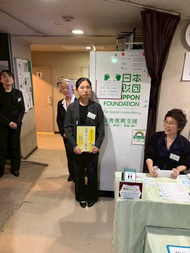 黒いジャケットを着たじゅん劇団員のなかむらあやかが「字幕サポートご案内係」と書かれた黄色い案内板を両手で持ち、受付け台のある会場入口で立って待機している様子。