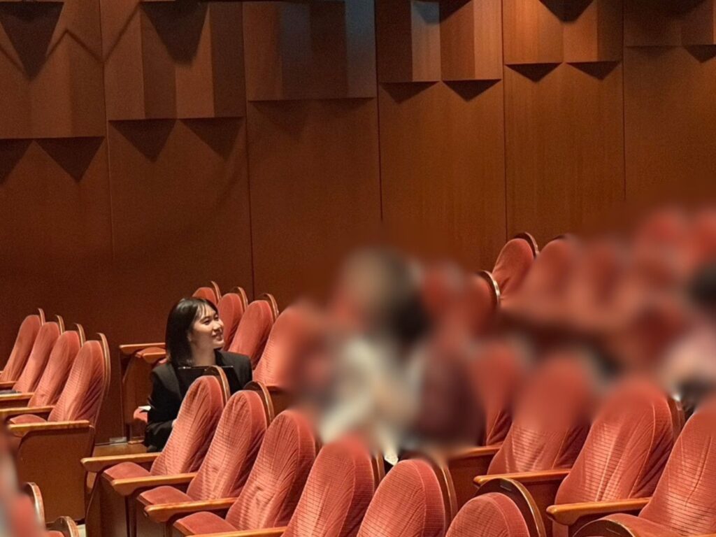 木目の壁と赤い座席が並ぶ劇場内で、黒いジャケットを着たかのうのぞみが客席のシートまえにしゃがみ、笑顔で斜め前方のりようしゃと話をしている。周囲には複数の赤い客席が配置されている。