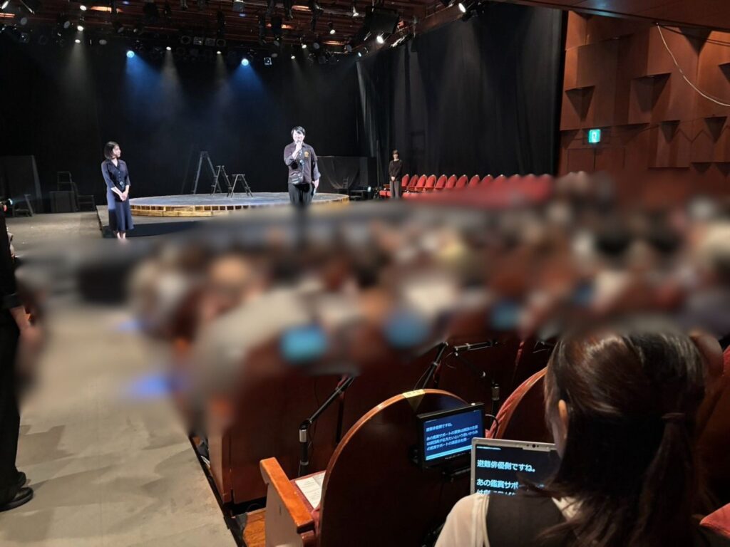 劇場内の客席後方からの風景で、手前では客席に座るこんどうまりあが字幕が表示されたモニターやタブレット画面を見つめている。前方奥の照明が当たった舞台上では、黒い服を着てマイクを持つおのあきひろと、こん色の服を着たかのうのぞみが立って観客に向かって話をしている。
