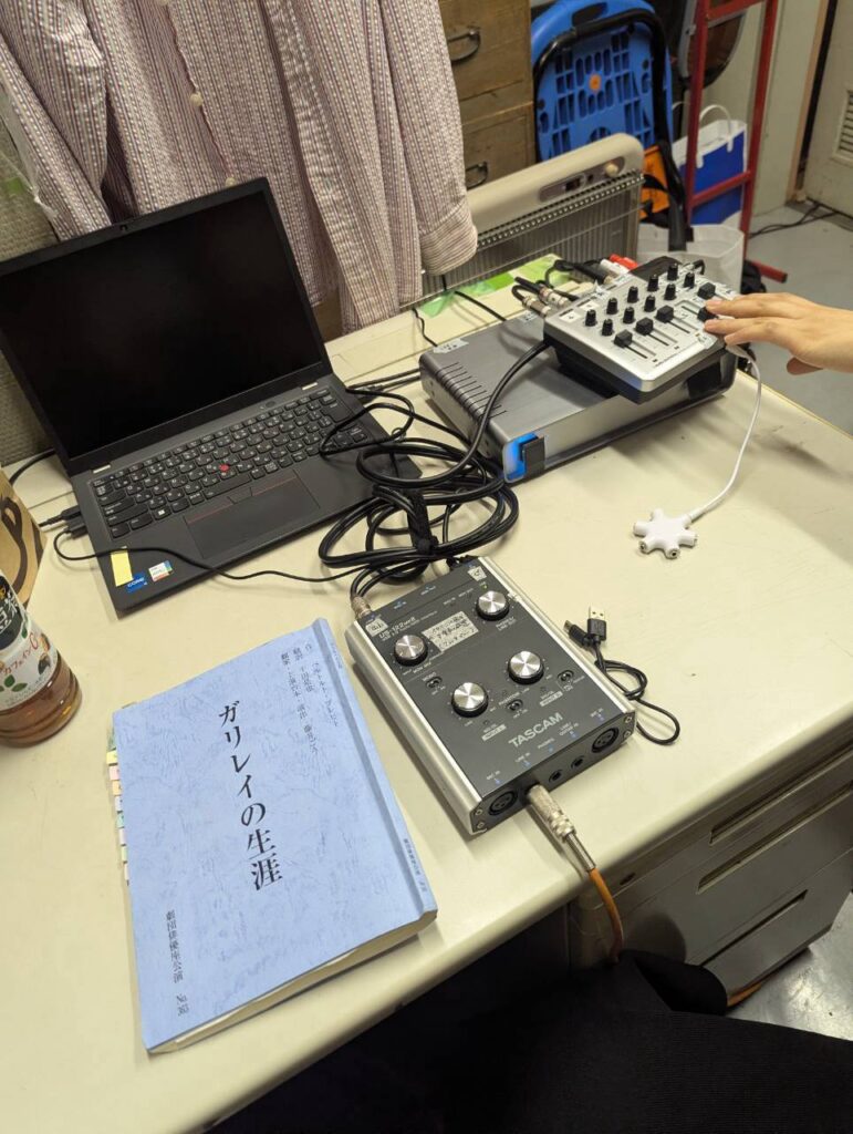 デスクの上に「ガリレイの生涯」と書かれた水色の台本、黒のノートパソコン、タスカムせいのオーディオインターフェイス、小さなミキサー機器が配置されている。人物の手が右上から伸びてミキサーのつまみを操作しており、機器同士は複数のケーブルで接続されている。