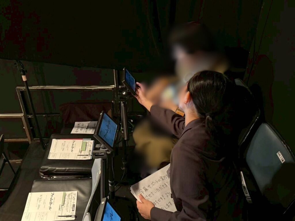 はいゆうざスタジオの客席で、黒い上着を着たなかむらあやかが、隣にいるりようしゃと一緒に、三脚に固定された小型モニターを見つめている。なかむらの手前や奥の席には、字幕サポートの案内やアンケート用紙などの書類が置かれ、別のモニターも設置されている。